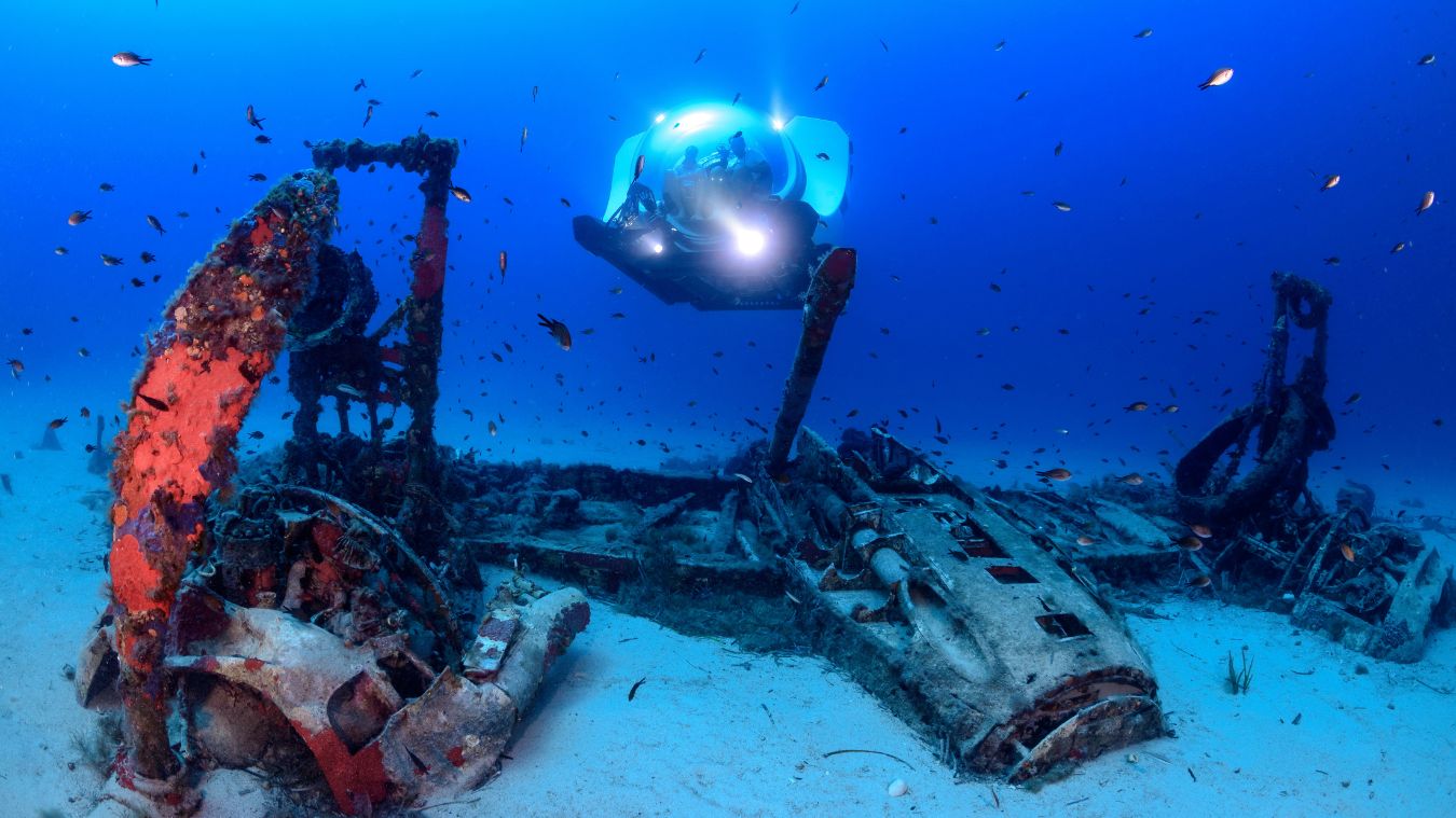 A deep-sea scene featuring the wreckage of a sunken ship, with rusted machinery and coral growths on the remains. A submersible with bright lights is exploring the wreck, surrounded by small fish swimming in clear blue water.