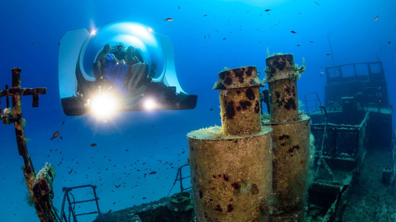 A submersible explores a shipwreck underwater, revealing coral-covered structures and marine life in a vibrant blue ocean.