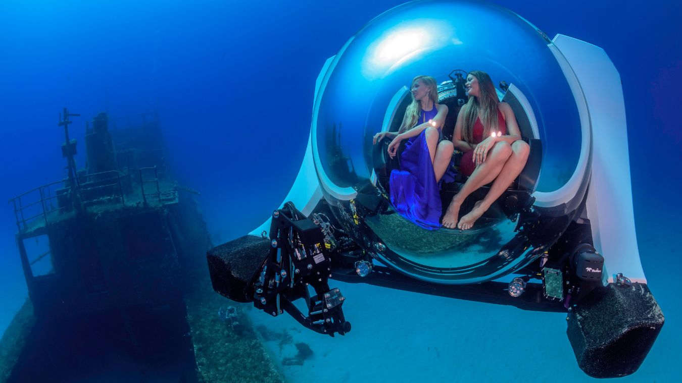Two women sit inside a sleek, futuristic submersible, exploring an underwater scene with a shipwreck in the background. The submersible's large, transparent dome allows for a panoramic view of the vibrant blue ocean.