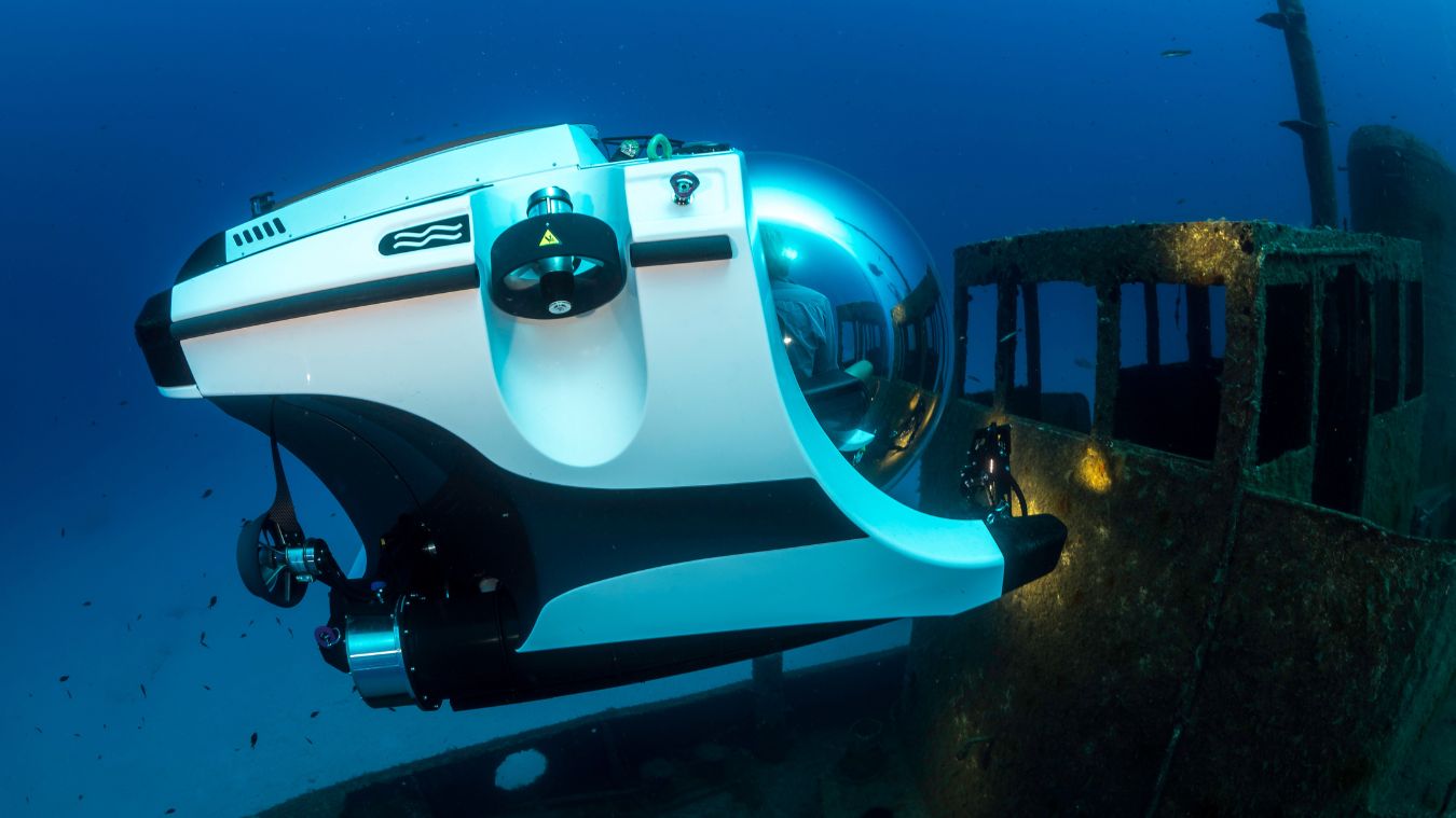 A submersible exploring the wreckage of a sunken ship underwater. The submersible features a sleek design with a glass dome and is surrounded by various fish in the blue sea.