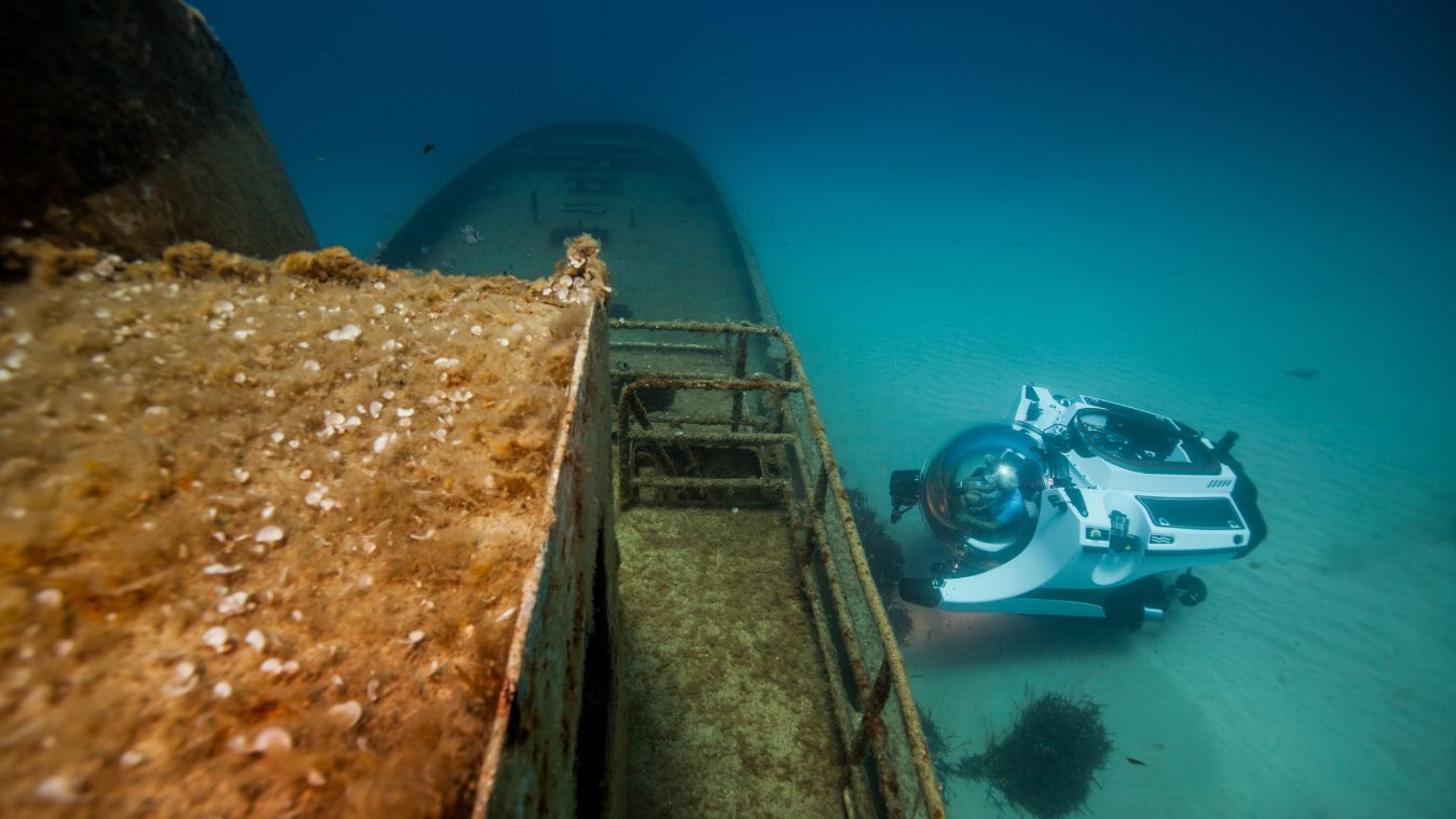 A submersible vehicle explores the wreck of a sunken ship, surrounded by clear blue water and various marine life.
