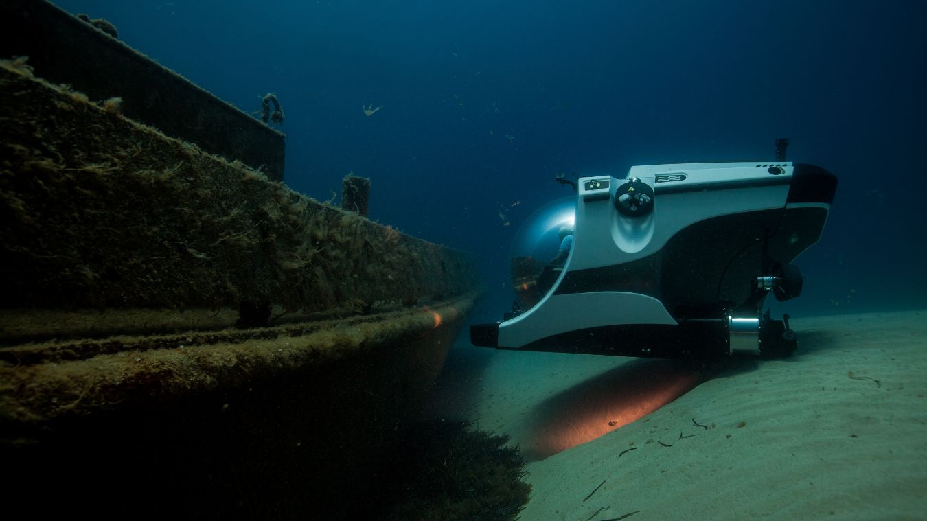 A small submersible exploring a shipwreck on the ocean floor, with sunlight illuminating the surrounding sand and debris.