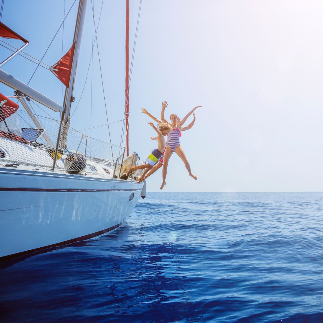 Two children joyfully leap from the side of a sailboat into bright blue ocean water, with the sun shining brightly overhead and red sail flags visible on the boat.