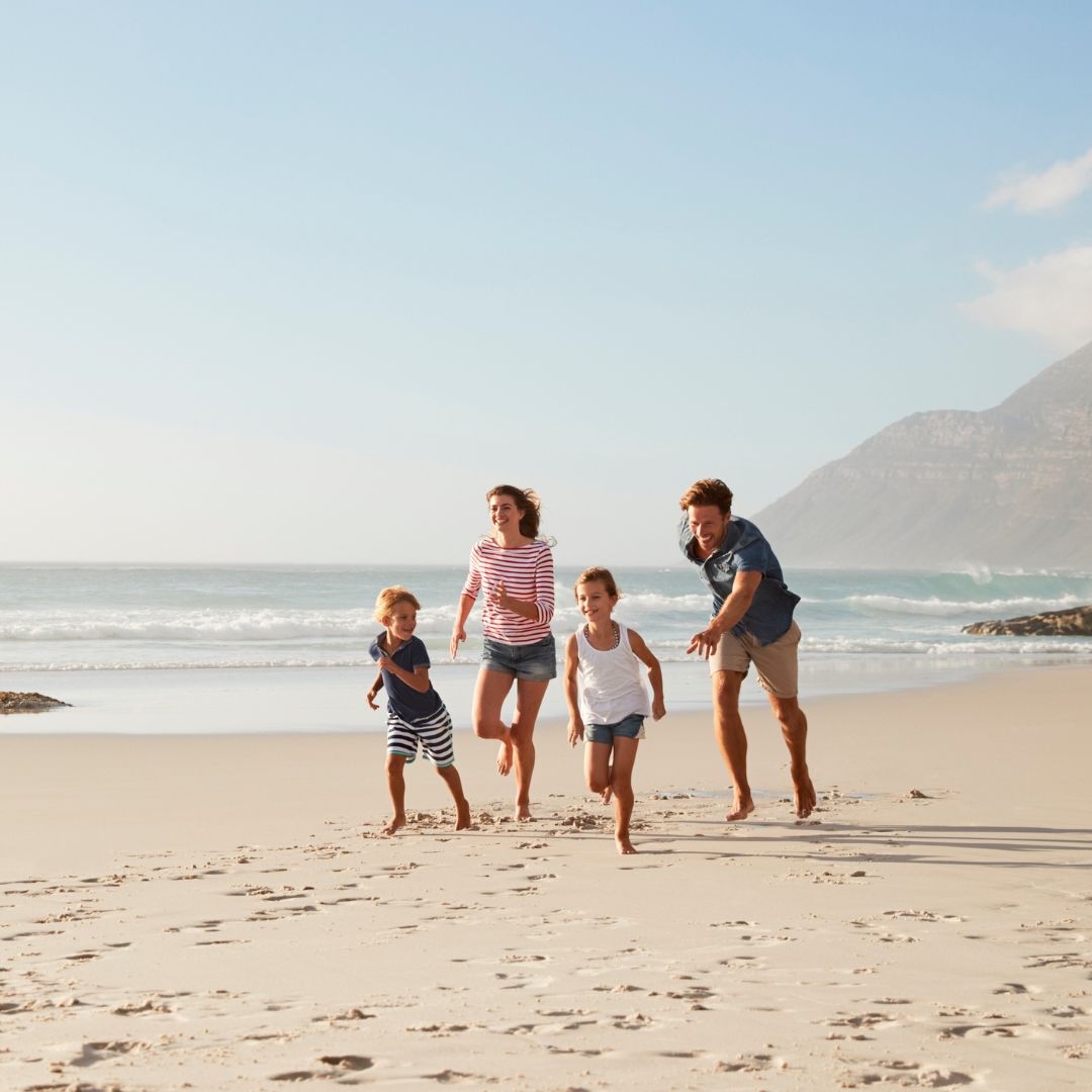 A family of four, including two children, joyfully runs along a sandy beach with waves crashing in the background. The sun shines brightly, casting a warm glow on the scene.