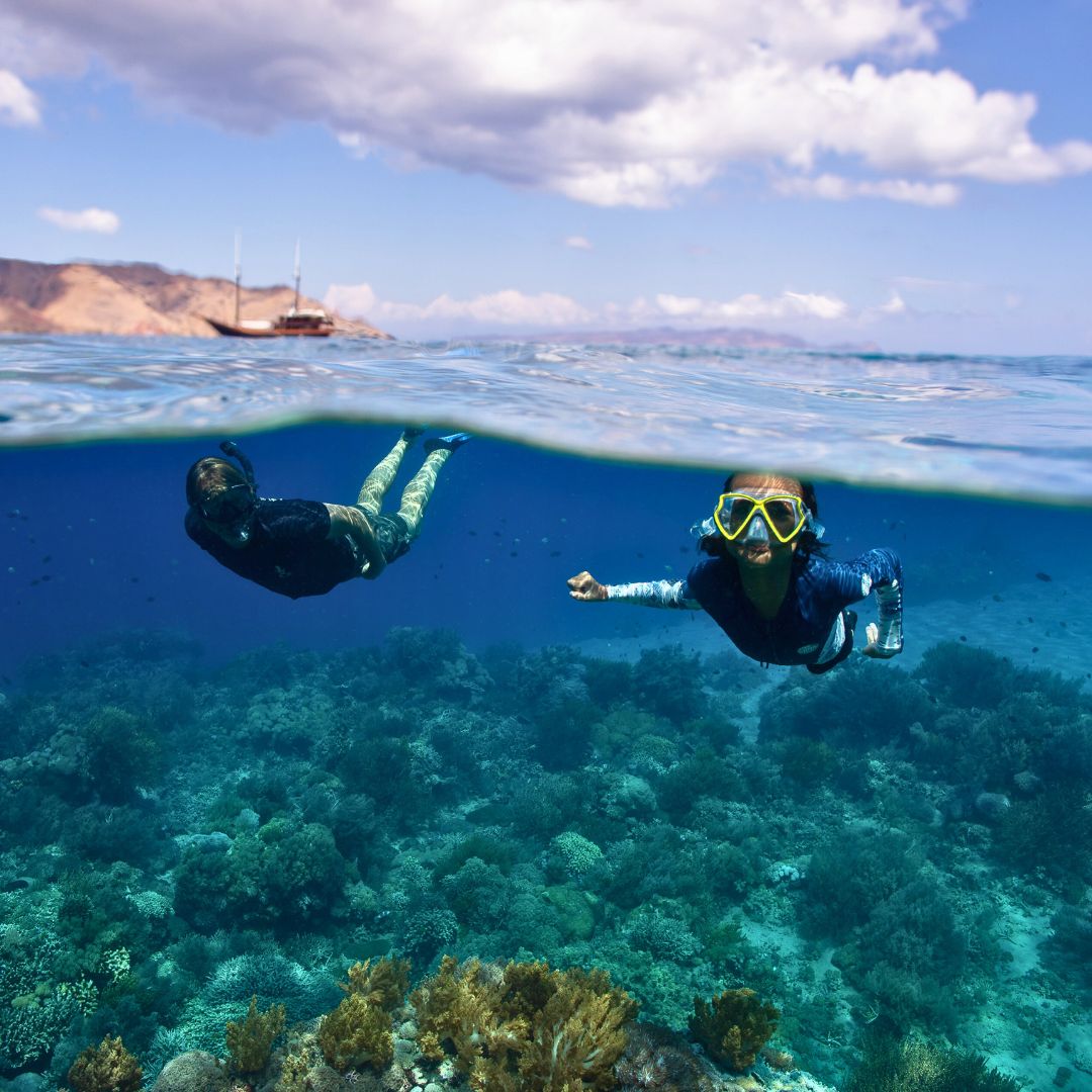 Two snorkelers swim in clear blue water above a vibrant coral reef. One snorkeler floats on the surface, while the other dives slightly deeper. A boat can be seen in the background against a backdrop of mountains and a cloudy sky.