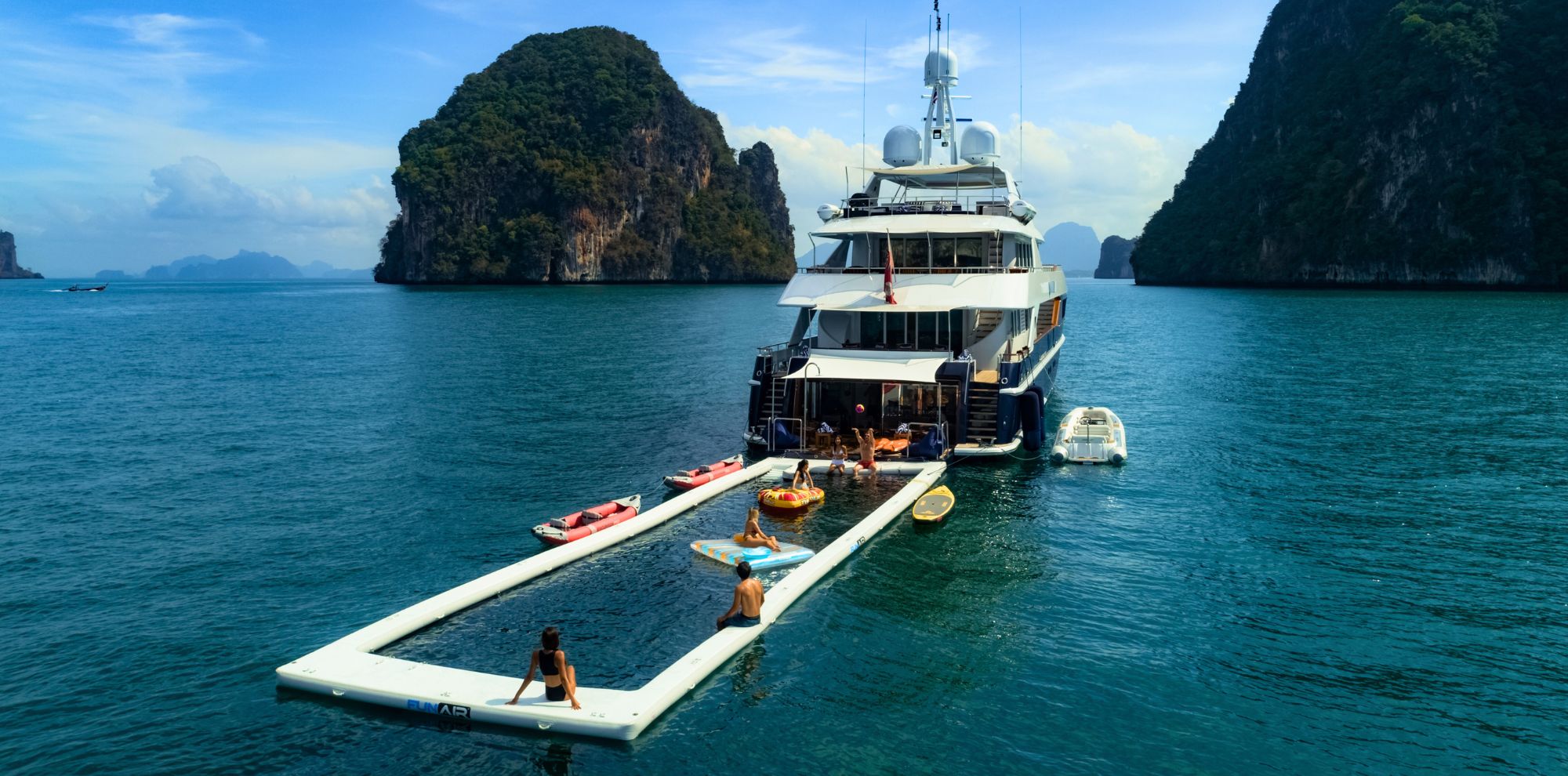 A luxury yacht anchored in calm turquoise waters, with several people enjoying various water activities. Surrounding limestone cliffs rise steeply in the background under a clear blue sky. A floating platform extends from the yacht, where some individuals are swimming and relaxing.