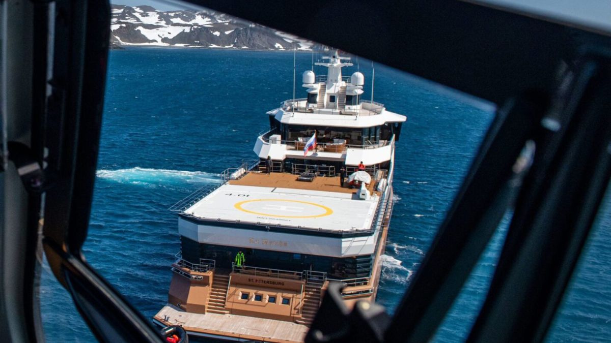 View from a helicopter window of a large expedition yacht with a helipad on the aft deck, anchored in deep blue arctic waters.