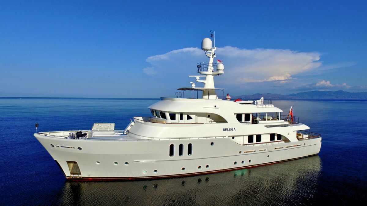 A large luxury yacht named "Beluga" is anchored in calm blue waters under a clear sky, with distant mountains visible on the horizon.