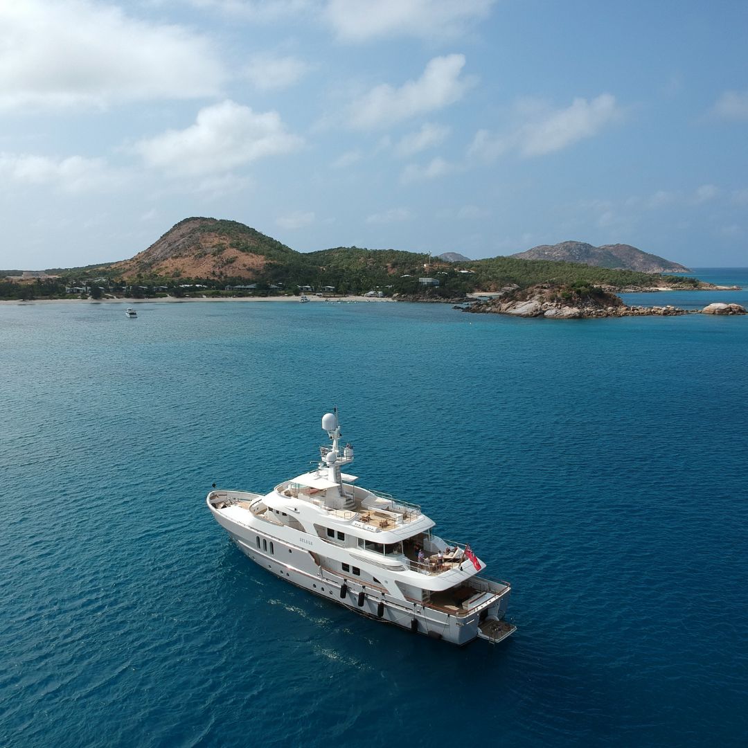 A luxurious yacht anchored in serene turquoise waters, with a backdrop of lush green hills and rocky islands under a partly cloudy sky.