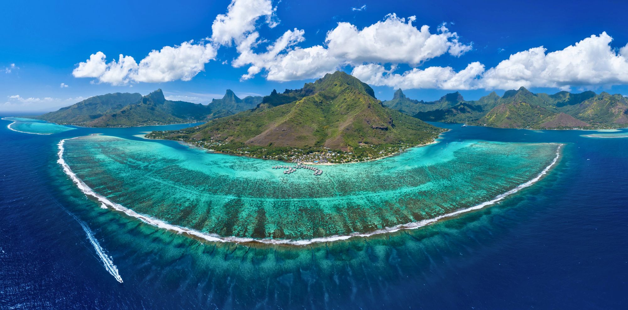 Aerial view of a stunning tropical island surrounded by vibrant turquoise lagoons and coral reefs. Lush green mountains rise in the background under a blue sky filled with white clouds.