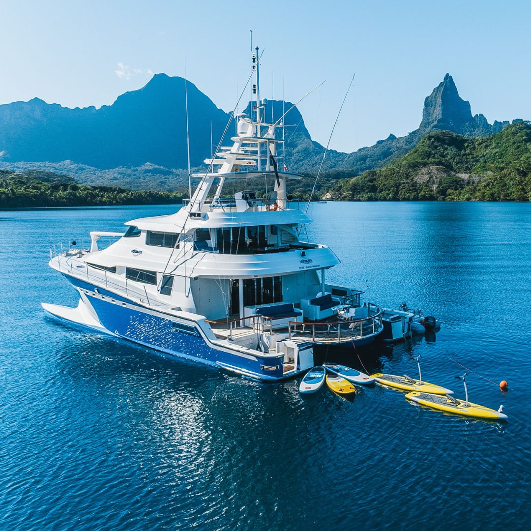 A large blue and white yacht anchored in calm blue waters, surrounded by lush green mountains under a clear blue sky. Three yellow kayaks are tied alongside the yacht.