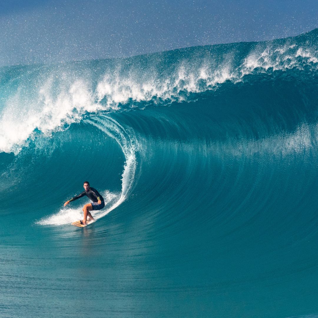 A surfer rides a large, turquoise wave, showcasing impressive skill and balance. The ocean is calm around the wave, creating a dramatic contrast.