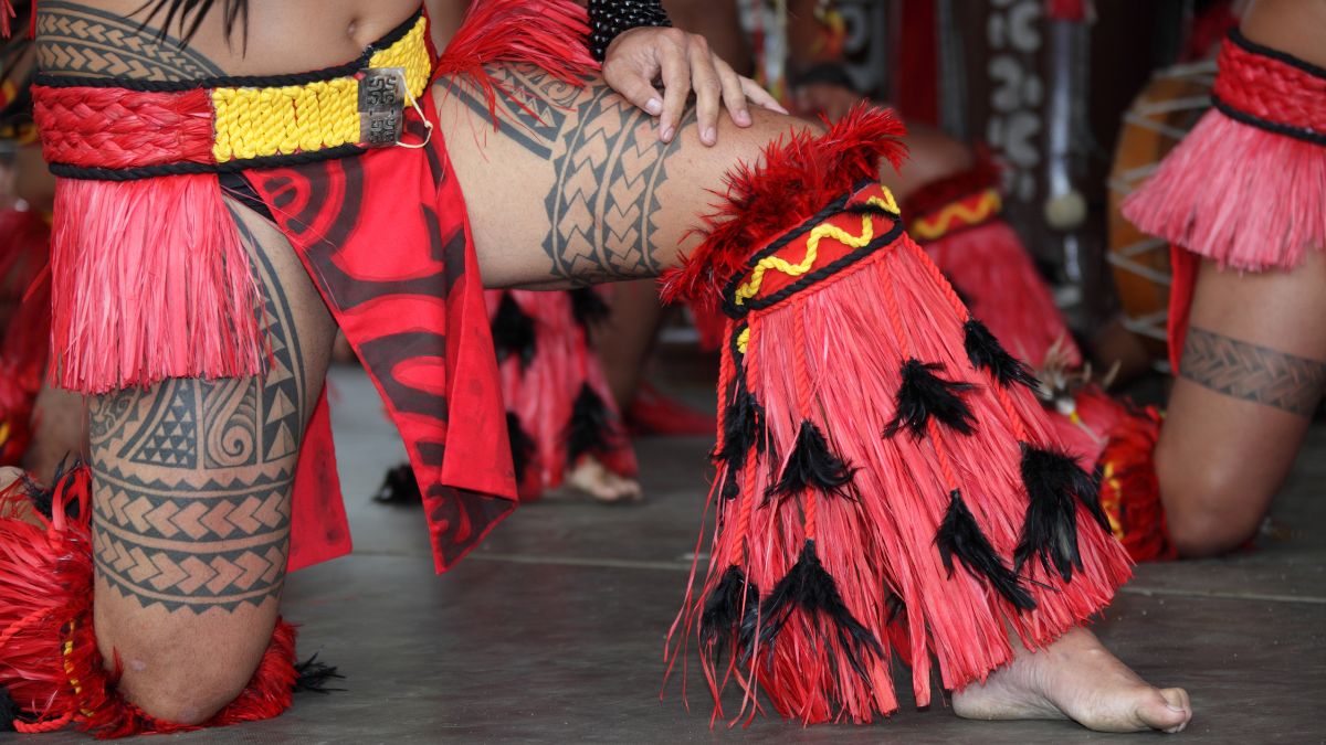 Traditional dancers display intricate tattoos and vibrant red outfits, featuring fringed skirts and decorative accessories. The focus is on their legs as they perform a cultural dance.