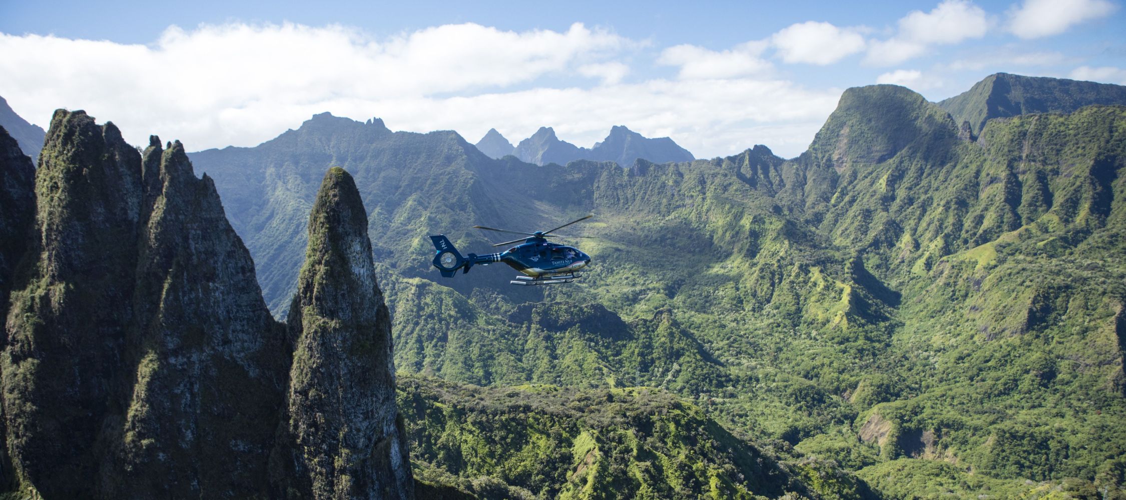 A helicopter flies amidst lush green mountains and rocky peaks under a partly cloudy sky.