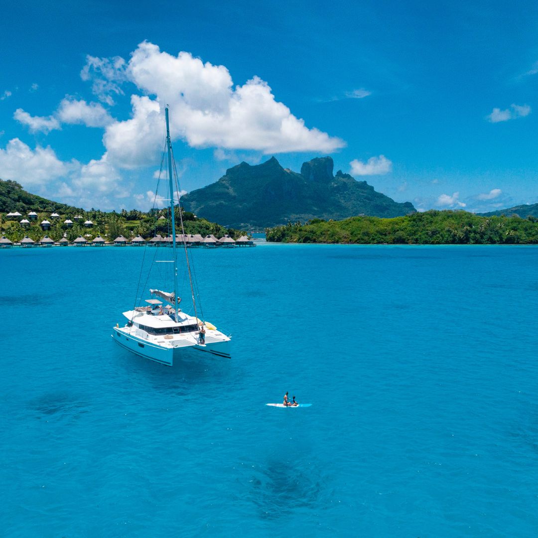 A sailboat floats on vibrant blue water near a lush green island with mountains in the background. A person paddleboards nearby under a bright blue sky with fluffy white clouds.