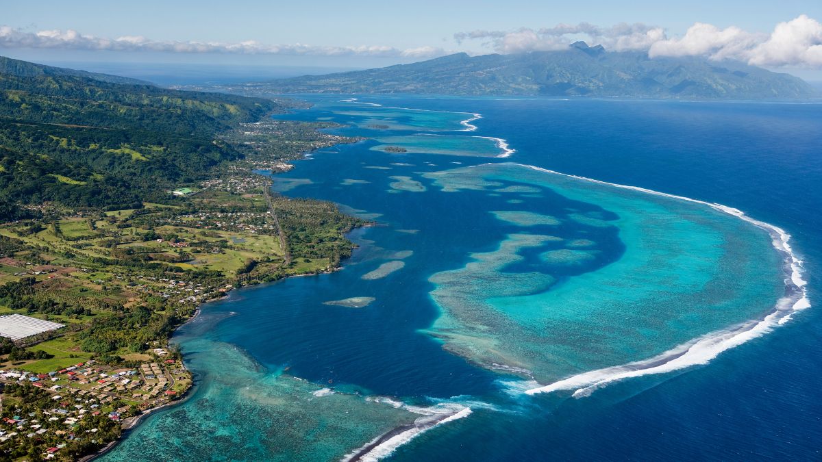 Aerial view of a vibrant blue lagoon bordered by a lush green coastline and distant mountains under a clear sky. Coral reefs are visible beneath the water, creating patterns in shades of blue and turquoise.