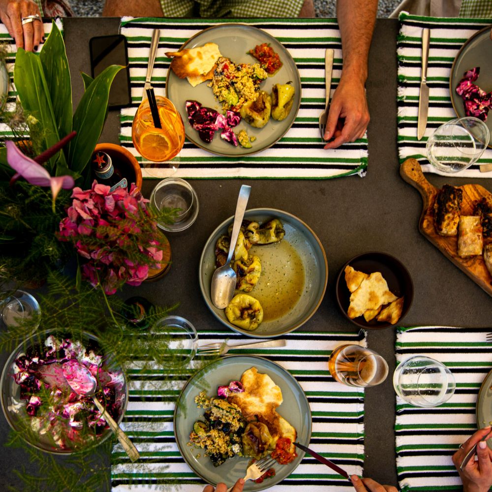 A top-down view of a dining table featuring colorful plates of food, including salads and dips, alongside drinks. Hands are reaching for the dishes, and vibrant flowers and greenery decorate the setting.