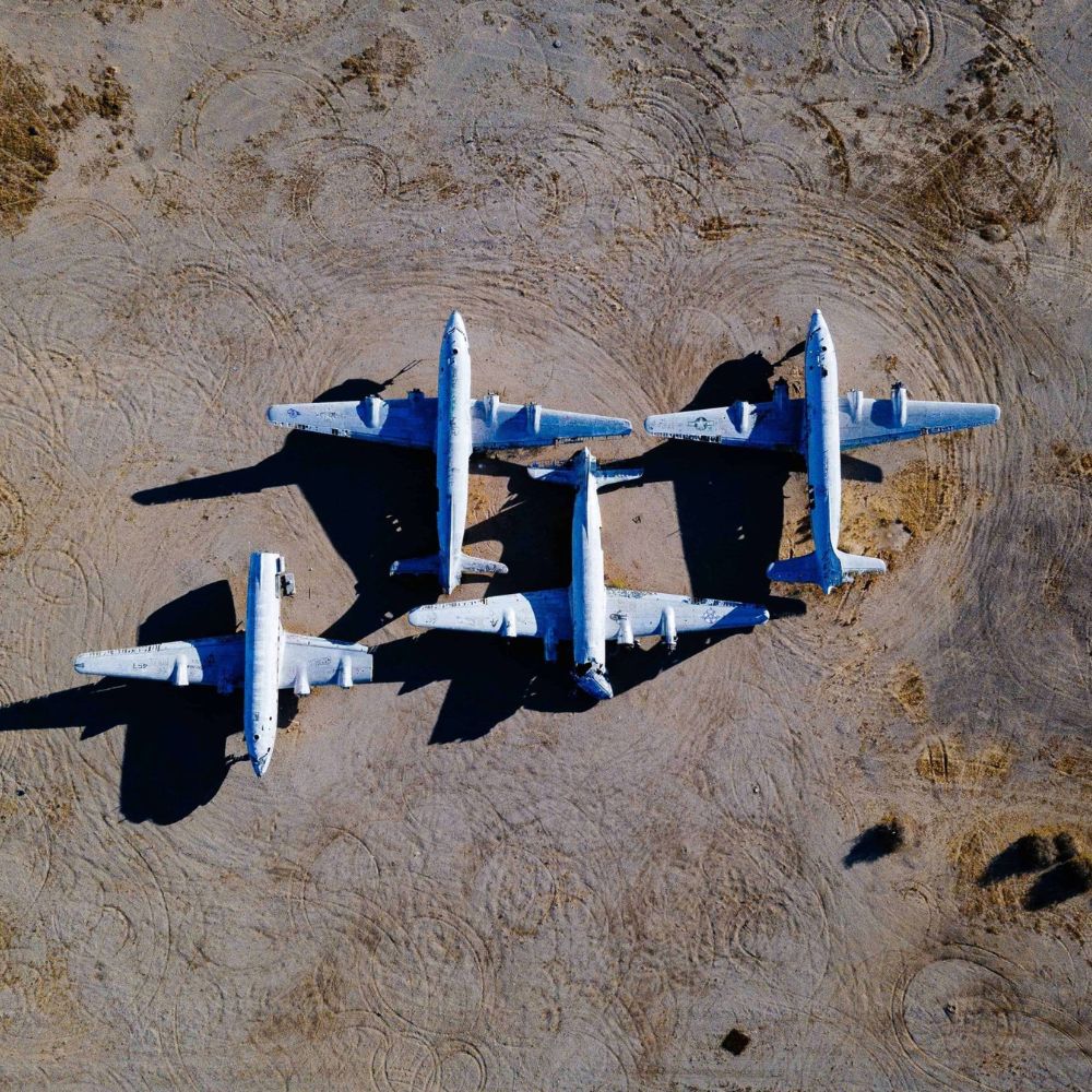 Four abandoned aircraft are arranged on a dirt field, casting distinct shadows. The planes display signs of wear and age, with peeling paint and rust. Circular tire marks are visible on the ground surrounding them.