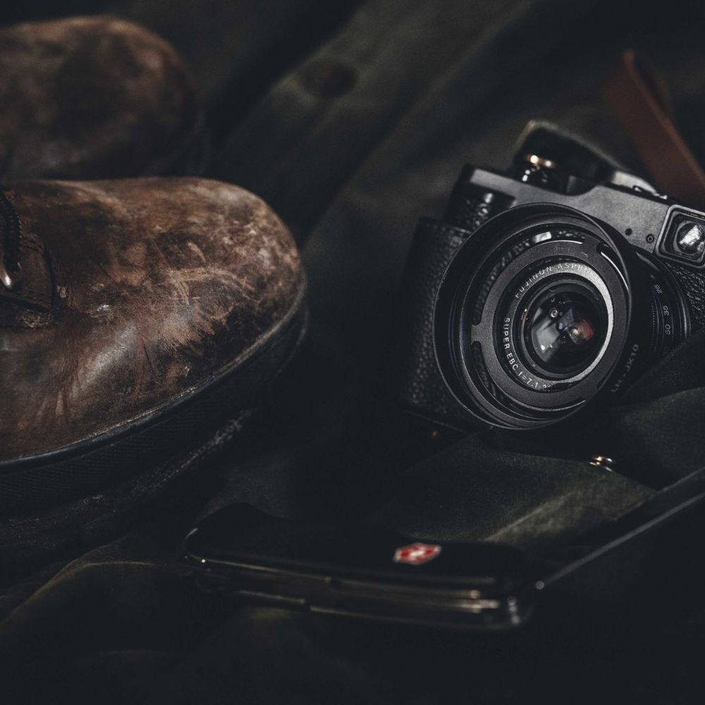 A pair of worn brown boots rests beside a camera and a black smartphone on a dark fabric surface.