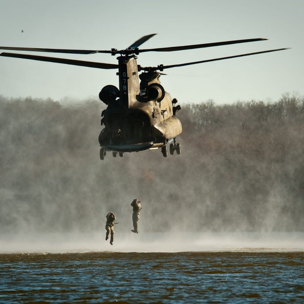 A military helicopter hovers above a body of water, with two soldiers rappelling down amid dusty clouds. The setting is outdoors, likely during a training exercise or operation.