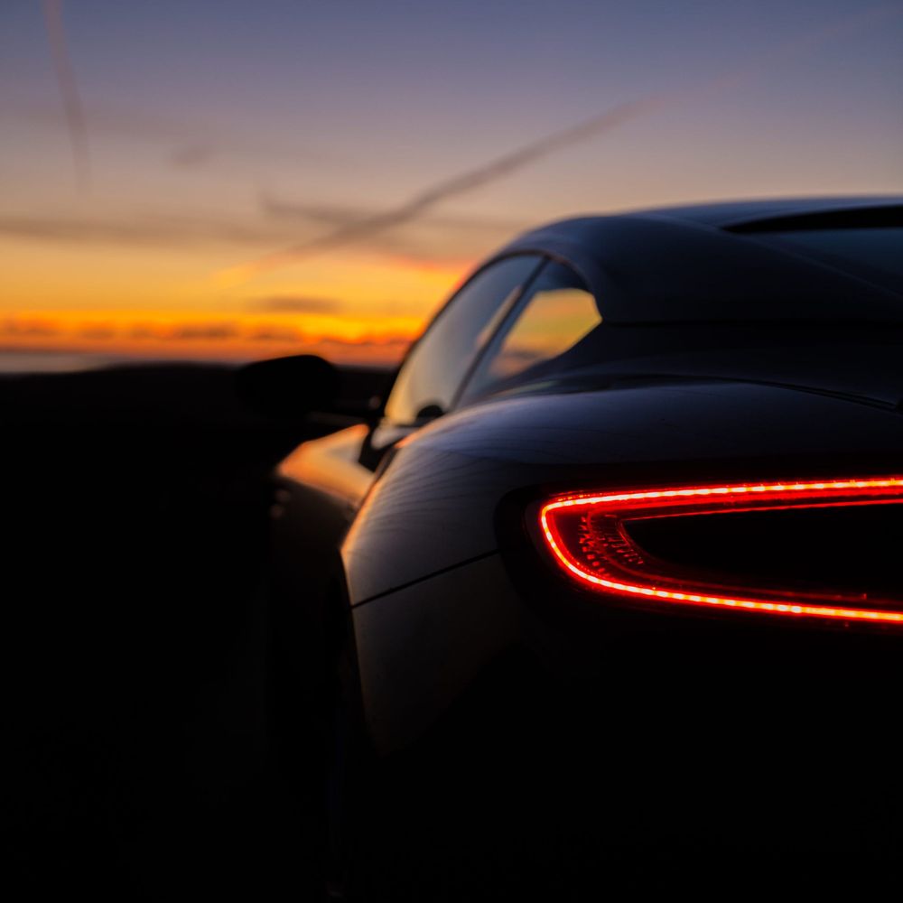 A sleek car with its rear light glowing, set against a vibrant sunset sky filled with shades of orange and purple.