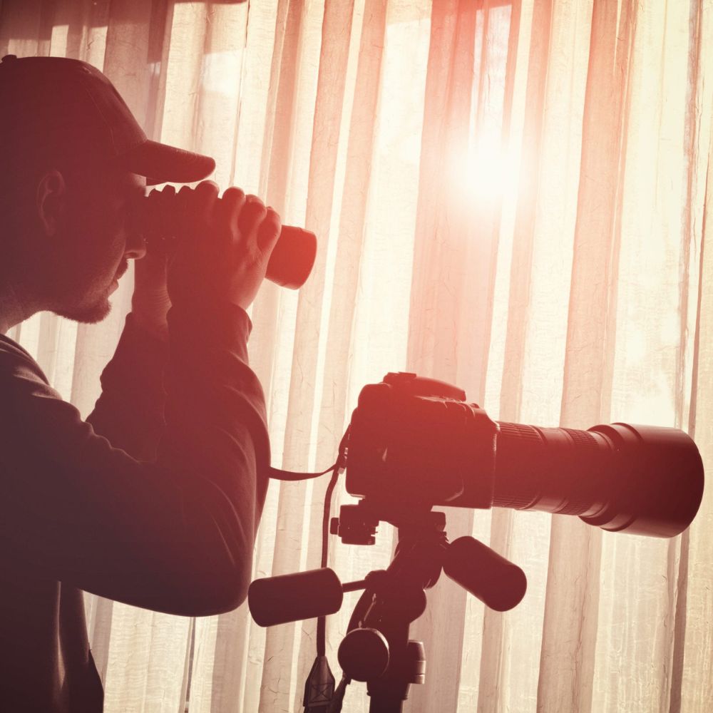 A person silhouetted against a window, looking through binoculars while a camera on a tripod stands beside them. Sunlight filters through sheer curtains, creating a warm glow.