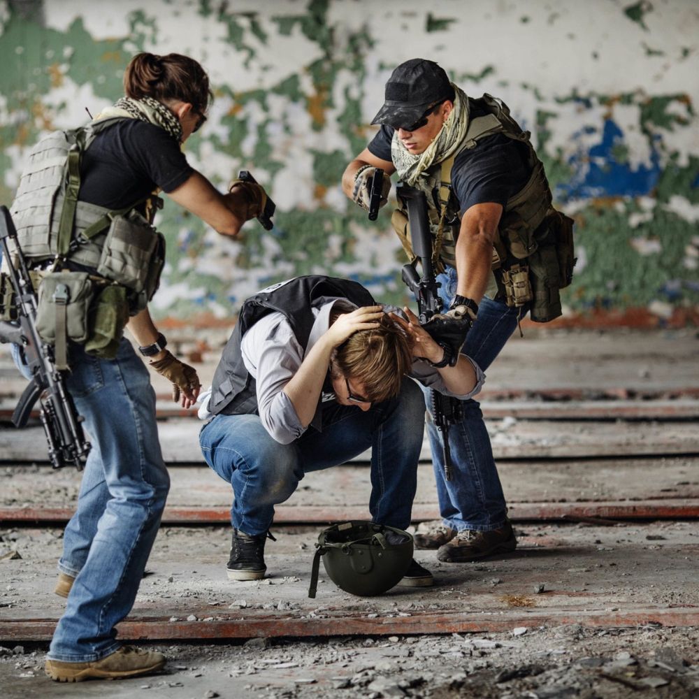 Two armed individuals assist a crouching person in a distressed position inside a dilapidated building. The environment appears gritty, with peeling paint and debris on the floor. The armed individuals wear tactical gear and face masks, suggesting a high-stakes situation.