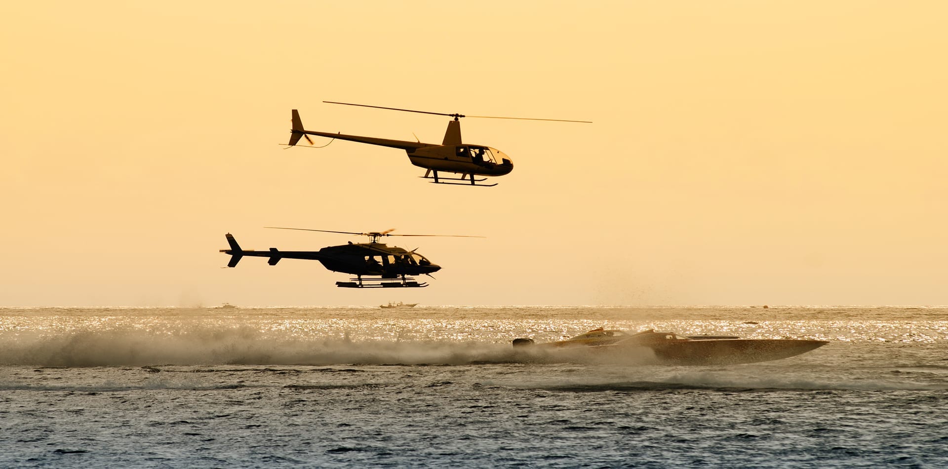 Two helicopters fly in the sky above a speeding motorboat, creating a dynamic scene against a golden sunset. The water sparkles with reflections from the setting sun.