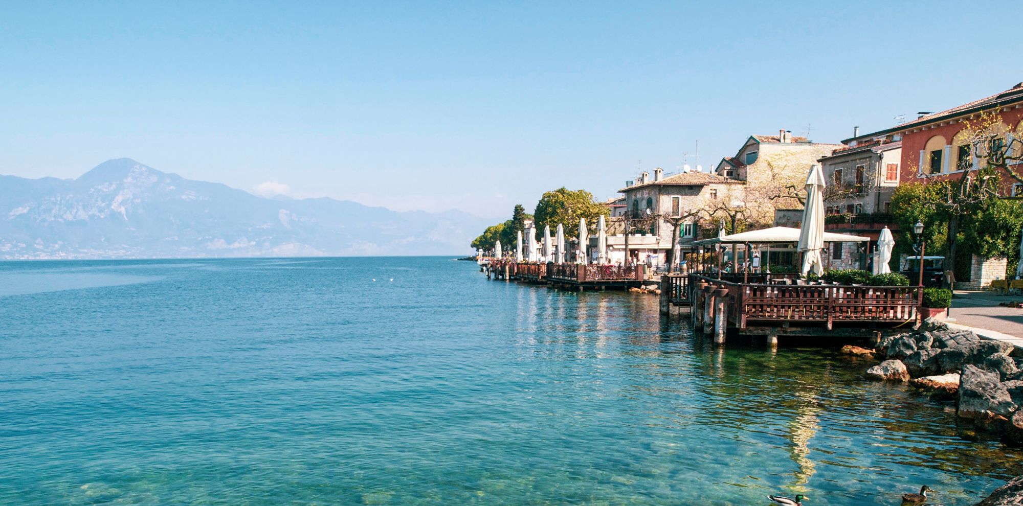 Outdoor cafe terraces on wooden piers overlook clear blue water and distant mountains under a bright sky.