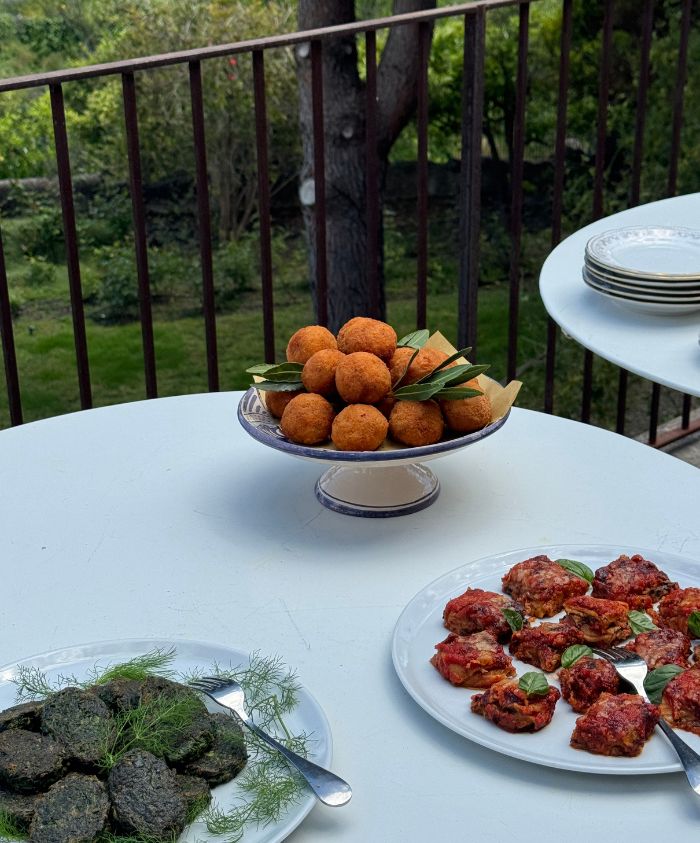 Three plates of appetizers, including arancini and small pastries, set on a white table on a terrace.