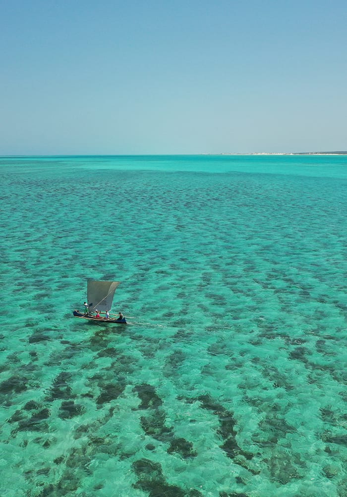 High-angle view of a small sailboat with a grey sail crossing vast, clear turquoise water with visible coral patterns.