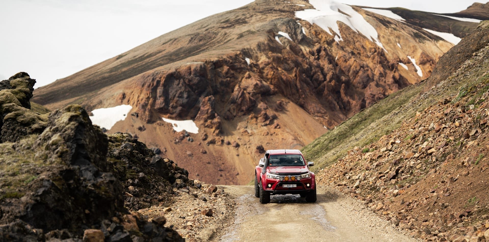A red off-road truck driving on a dirt path through a rugged mountain landscape with patches of snow.