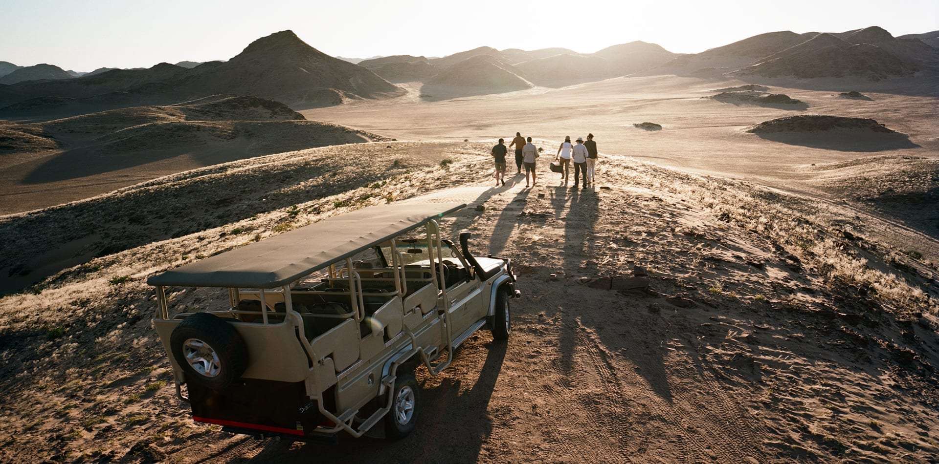 Group of people walking away from an open-sided safari vehicle toward a vast, hilly desert landscape at sunset.