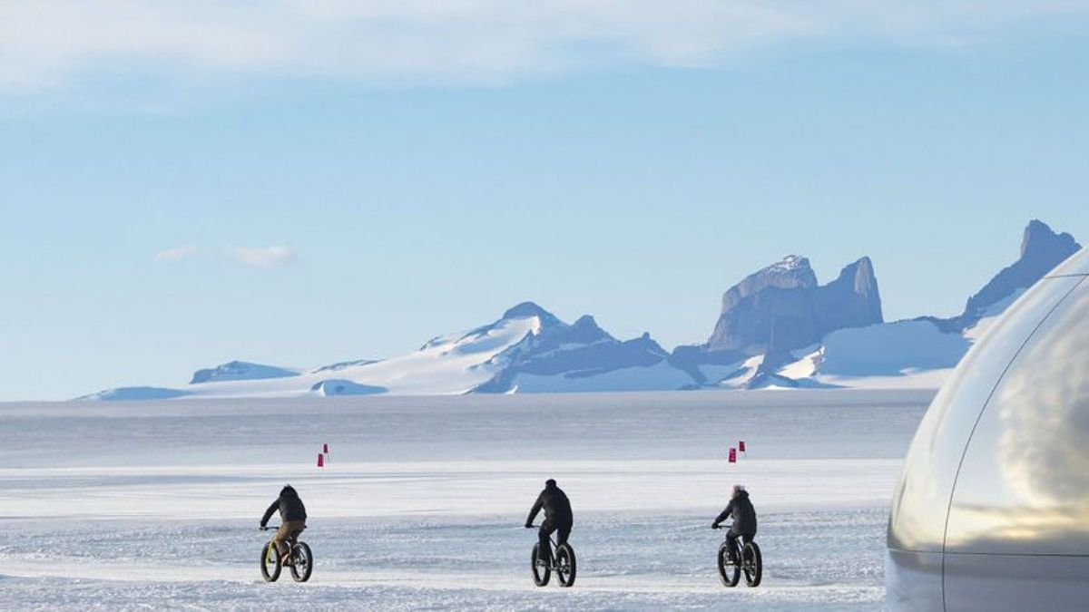Three cyclists ride fat bikes across a snowy landscape, with distant mountains and a clear blue sky in the background.