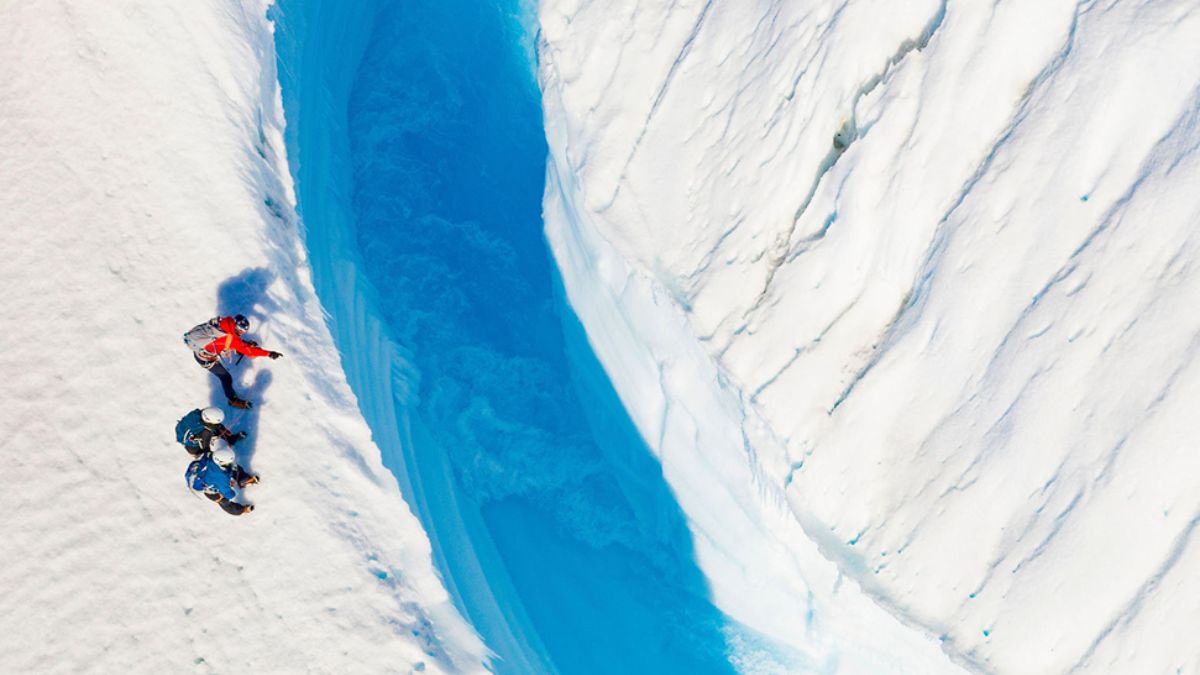 Three climbers explore a stark, icy landscape, standing near a deep blue crevasse in a glacier. The bright blue water contrasts with the surrounding white snow and ice formations.