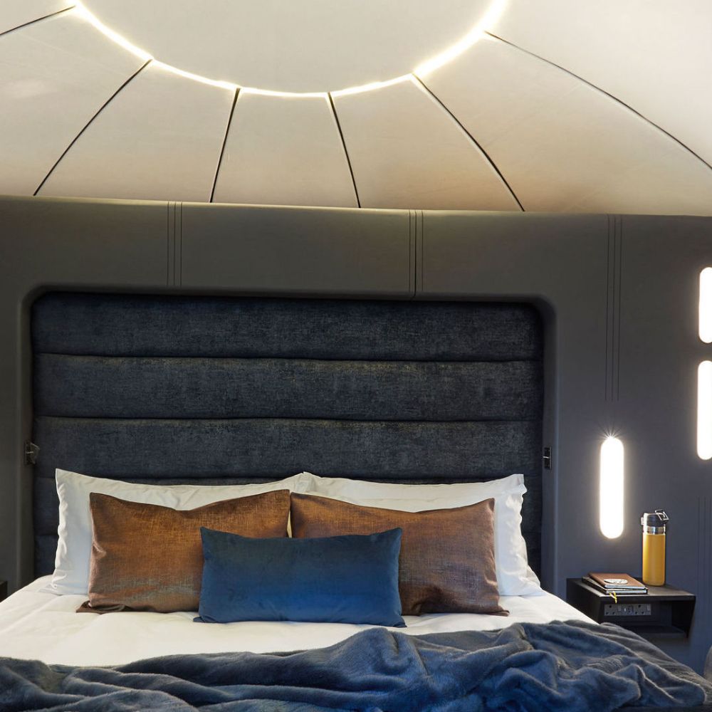 A modern yacht cabin featuring a plush bed with dark blue and bronze accents. The headboard has a sleek design, and warm lighting creates a cozy atmosphere. A small bedside table holds a glass of orange juice, complementing the luxurious decor.