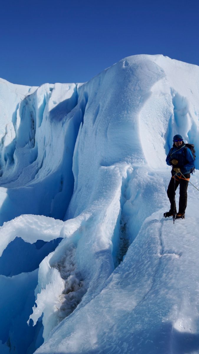 A climber stands on a jagged ice formation, surrounded by towering blue ice and deep crevasses under a clear blue sky.