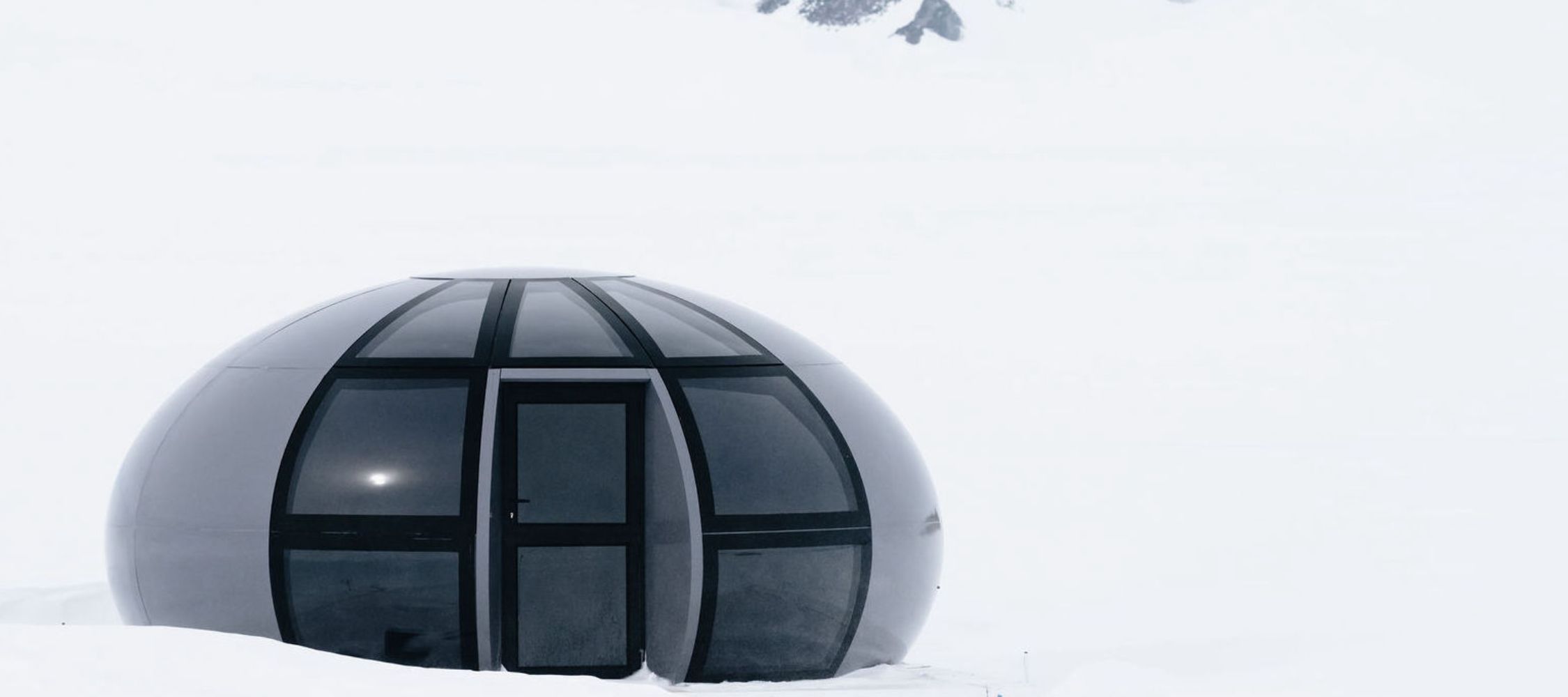 A futuristic, spherical black structure surrounded by a vast expanse of white snow, set against a backdrop of mountains.