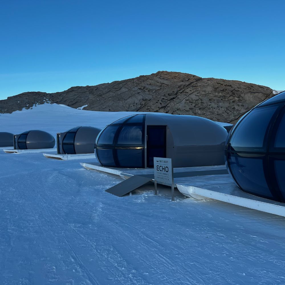 Translucent igloos on a snowy landscape with rocky hills. A sign reading 'ECHO' near the closest igloo. Clear sky with soft blue light
