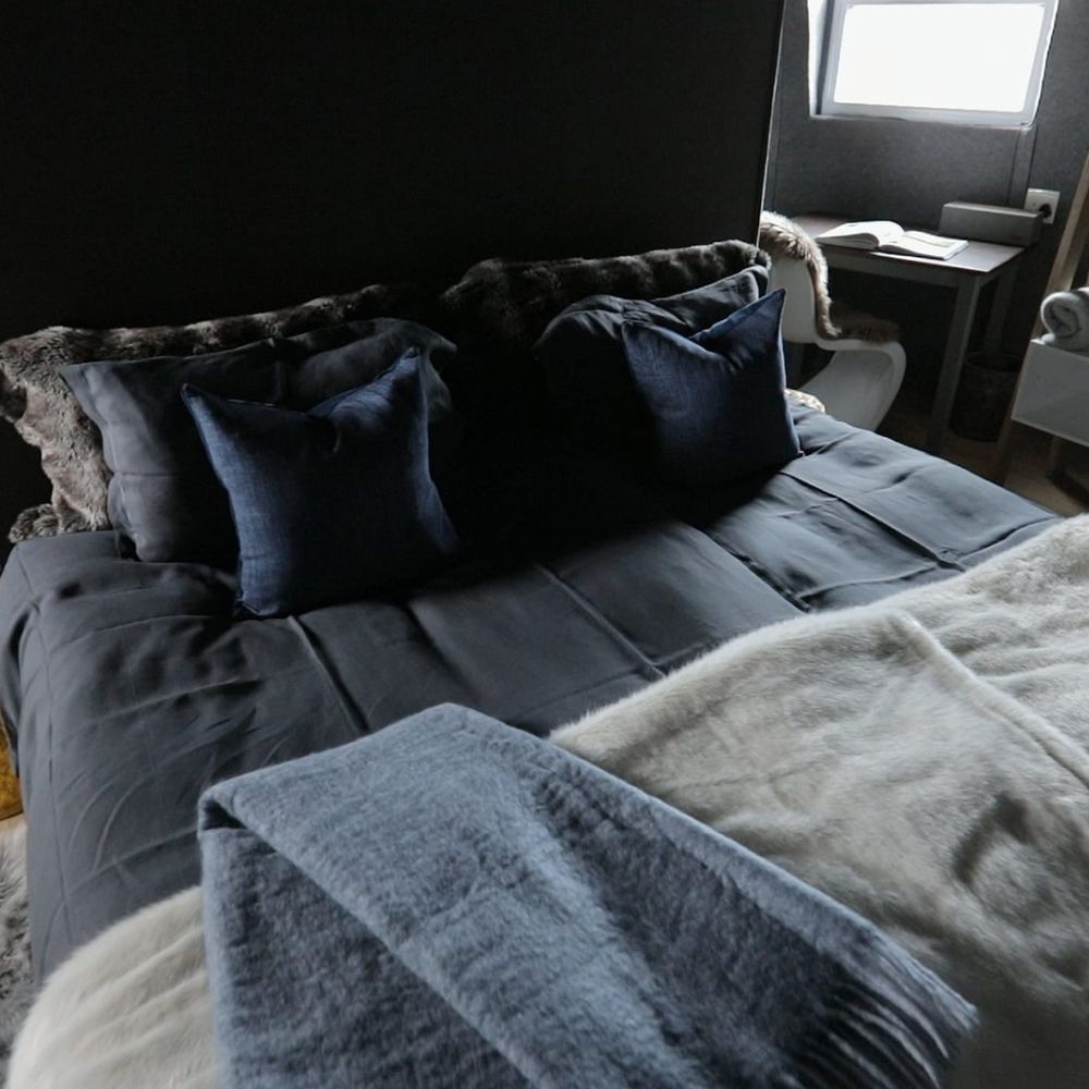 A cozy bed dressed in dark linens, featuring plush navy cushions and a soft, light-colored throw. The room is softly lit, with a minimalist desk and chair visible in the background.