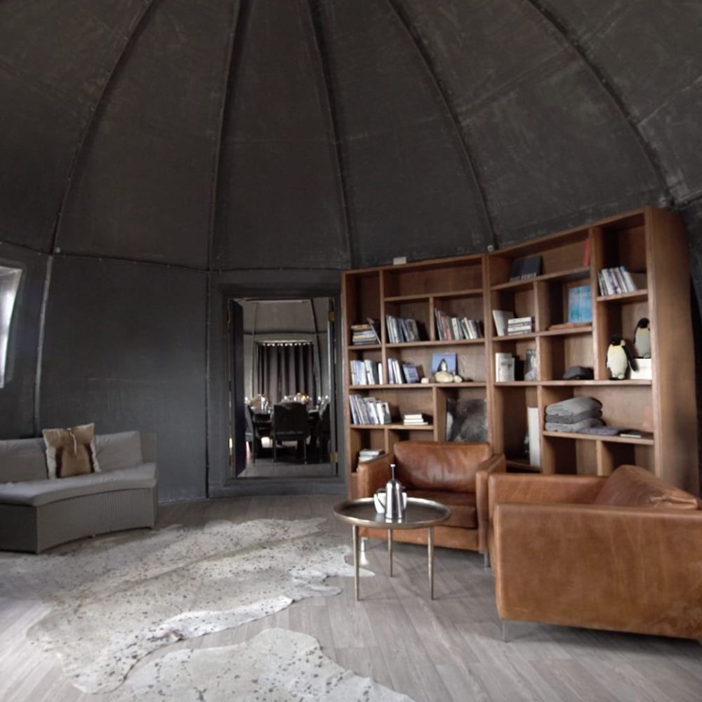 A stylish interior of a dome-shaped room featuring a cozy seating area with leather armchairs and a sofa, a wooden bookshelf filled with books, and a decorative rug on the floor. Natural light enters through windows, creating a warm atmosphere.