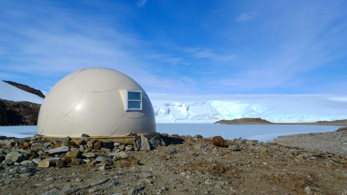 A small, beige dome-shaped structure sits on rocky terrain next to icy formations under a bright blue sky, with snow-capped mountains in the background.