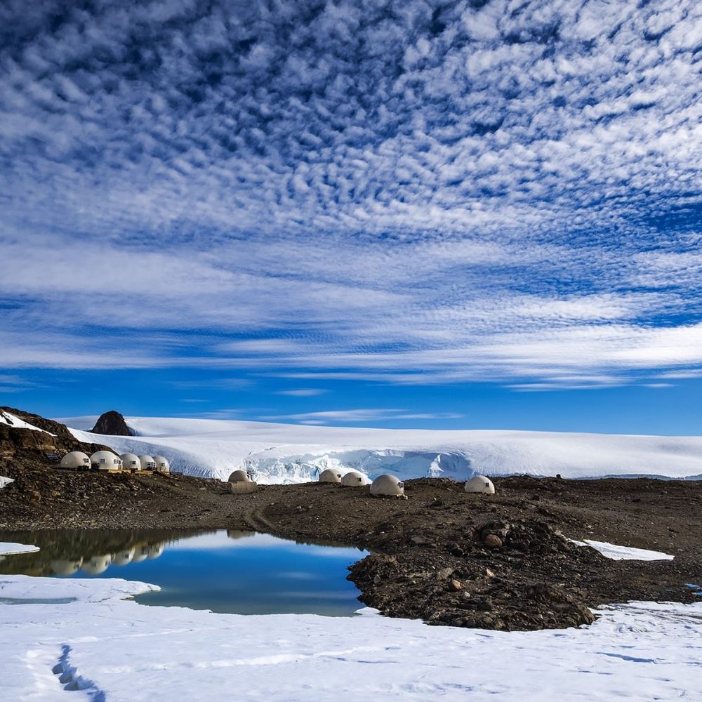 A remote landscape featuring white dome structures on rocky terrain, surrounded by patches of snow and a small body of water. In the background, a glacier is visible under a bright blue sky filled with wispy clouds.