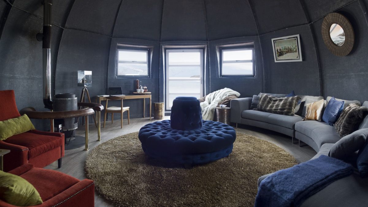 A modern, circular living room featuring a blue tufted ottoman in the centre, surrounded by a cozy grey sofa, red armchair, and a patterned rug. Large windows provide a view of the landscape outside. A small desk with a chair is positioned against one wall, and soft cushions and throws add warmth to the space.