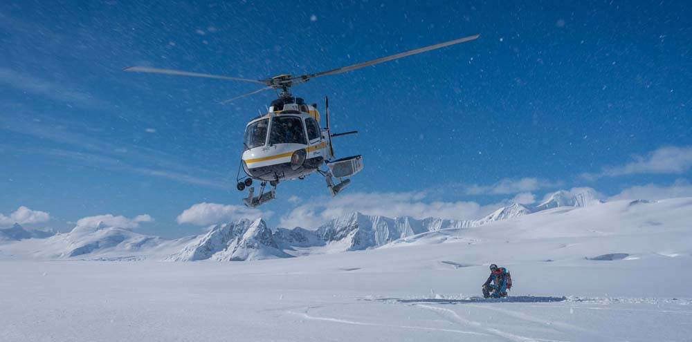 A helicopter hovers above a snowy landscape, with towering mountains in the background. Below, a person kneels on the snow, surrounded by pristine white terrain.