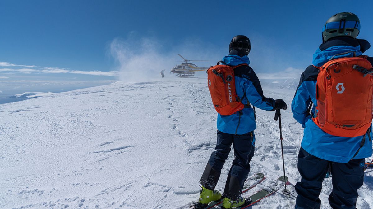 Two skiers heli skiing in antarctica in bright blue jackets and orange backpacks stand on a snowy mountain peak, looking towards a helicopter landing in the distance. The sky is clear and blue, with a light breeze creating a spray of snow.