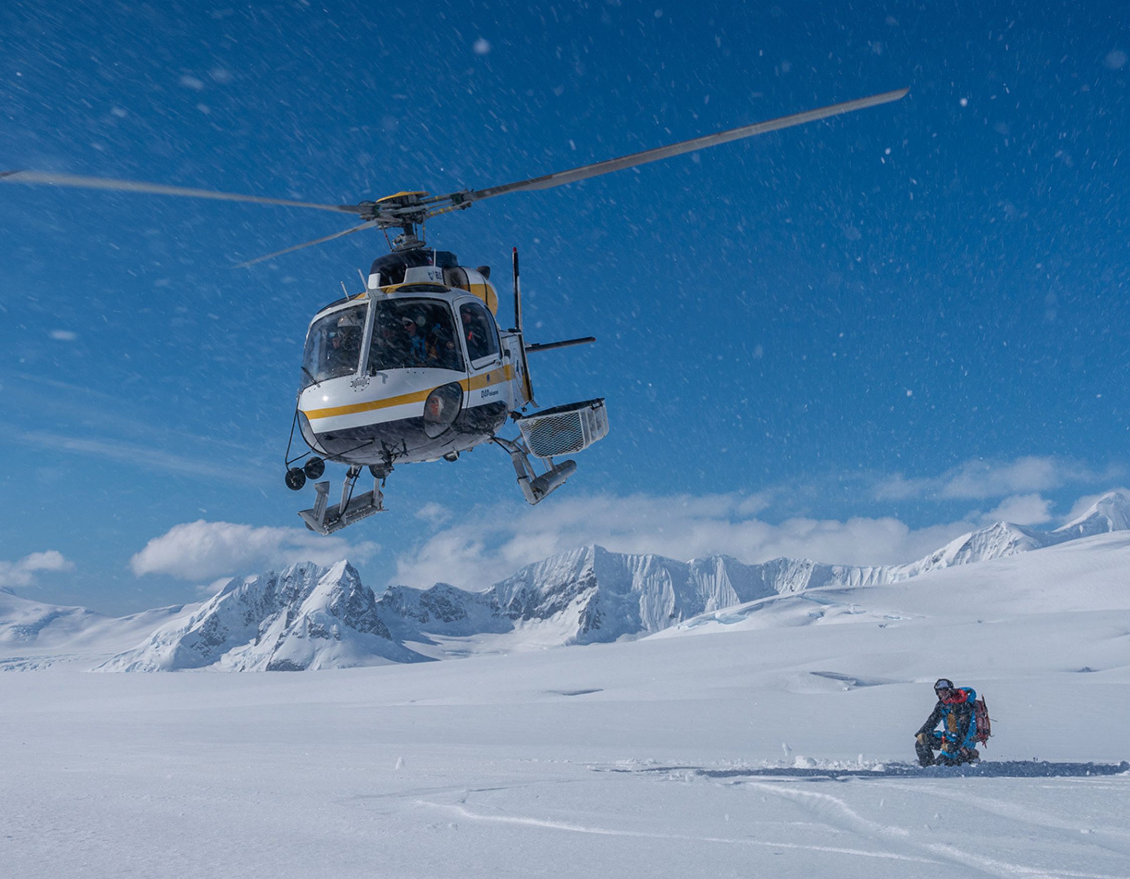 A helicopter hovers above a snowy landscape while a person kneels in the snow, surrounded by mountains under a clear blue sky. Snowflakes are visible in the air, creating a dynamic winter scene.