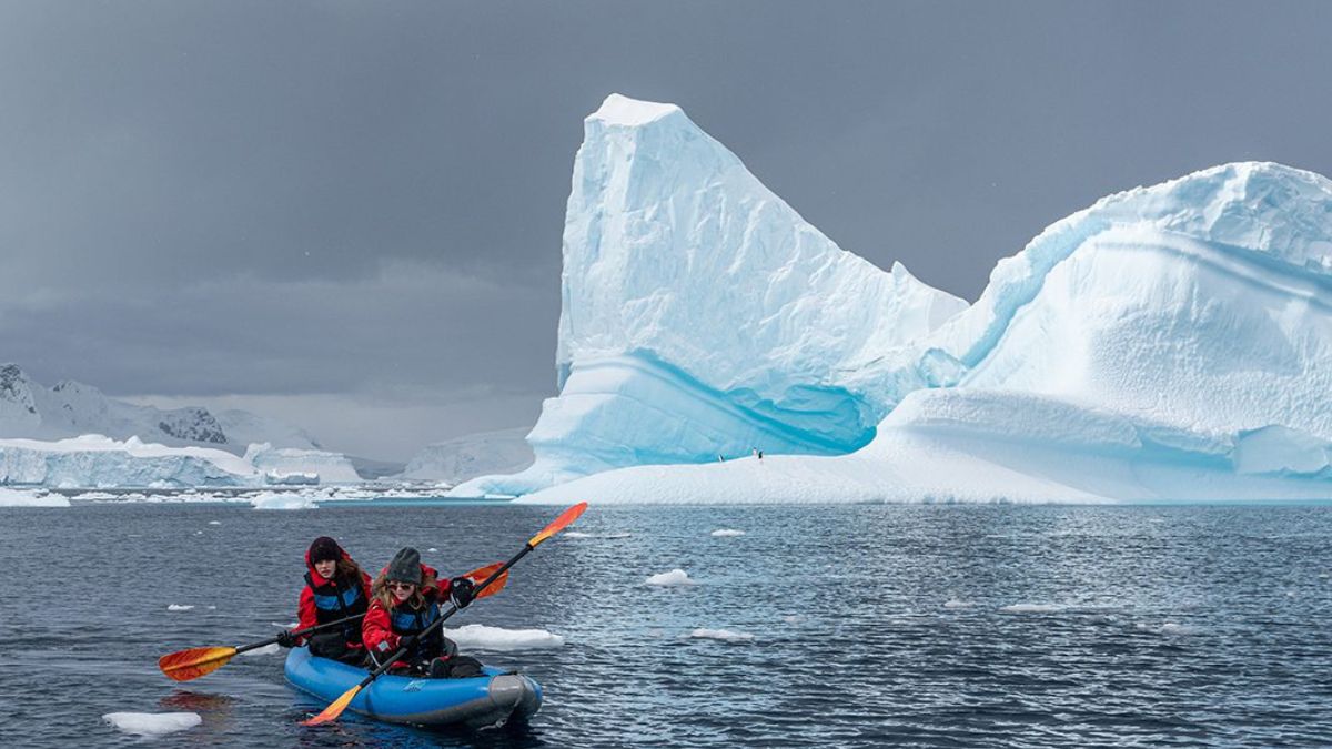 Two kayakers navigate icy waters with a large blue iceberg in the background. The dramatic sky is overcast, enhancing the surreal landscape.