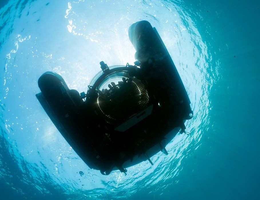 A submersible glides beneath the water's surface, casting a shadow with sunlight filtering through the clear blue sea above. Bubbles and marine life can be faintly seen around it.