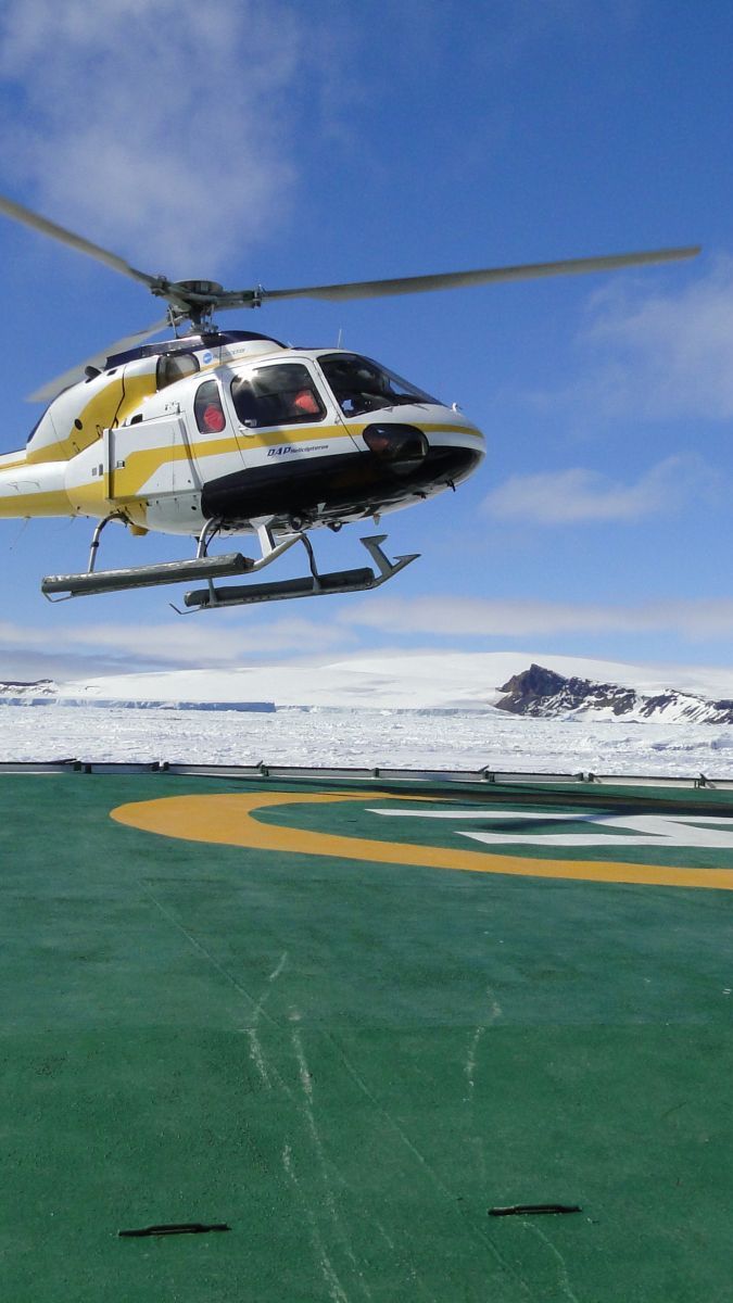A helicopter hovers above a green and yellow helipad with a snowy landscape and mountains in the background.